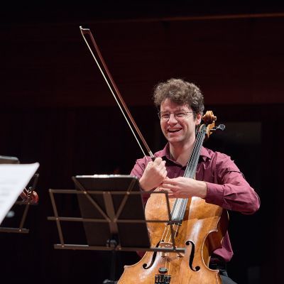 Male presenting student laughing on stage holding their cello whilst seated